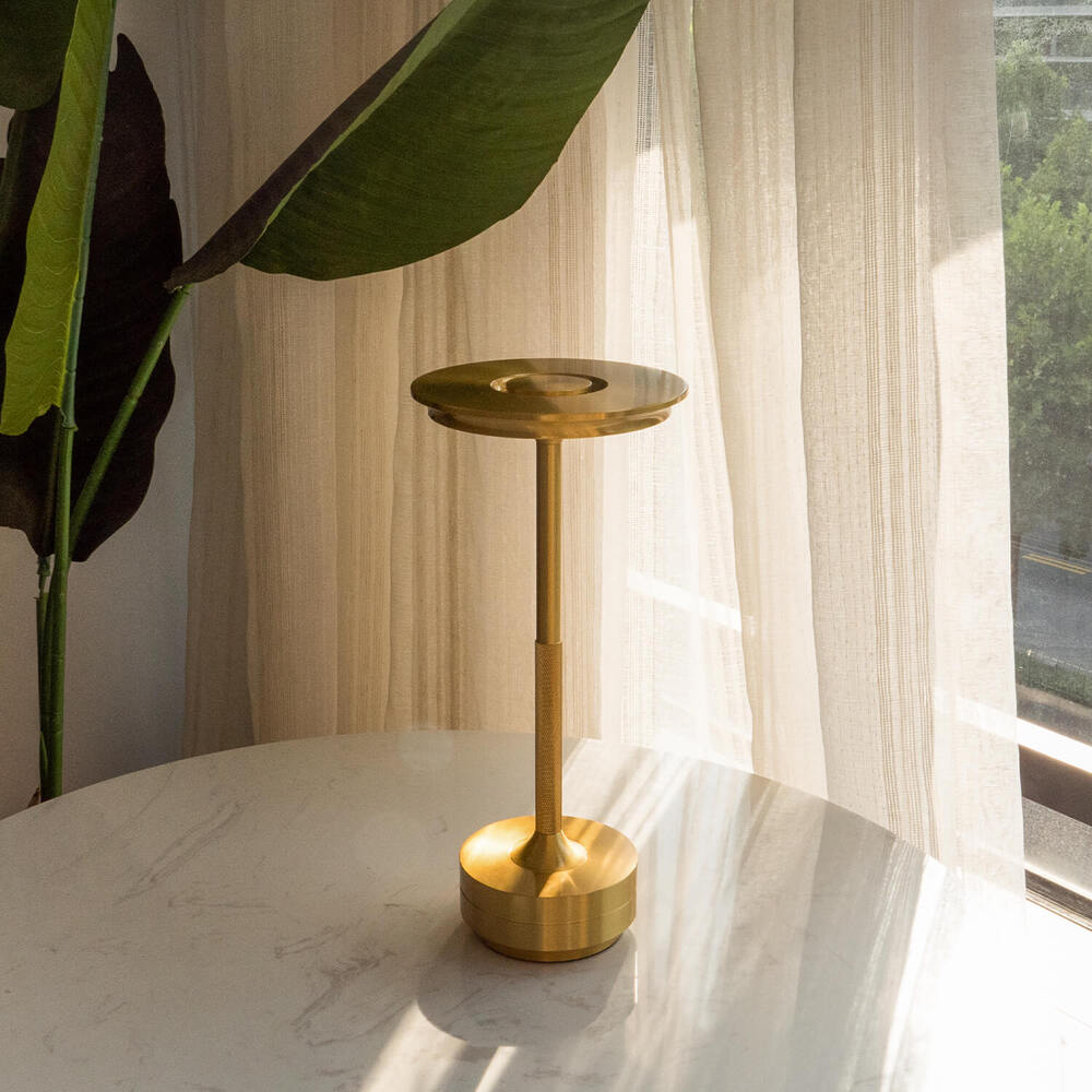 Sleek brass cordless table lamp on a white table near a large plant, bathed in natural sunlight.