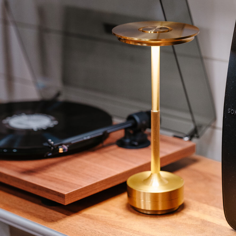 Sleek cordless table lamp with a brass finish next to a vinyl turntable, providing ambient lighting for a stylish setting.