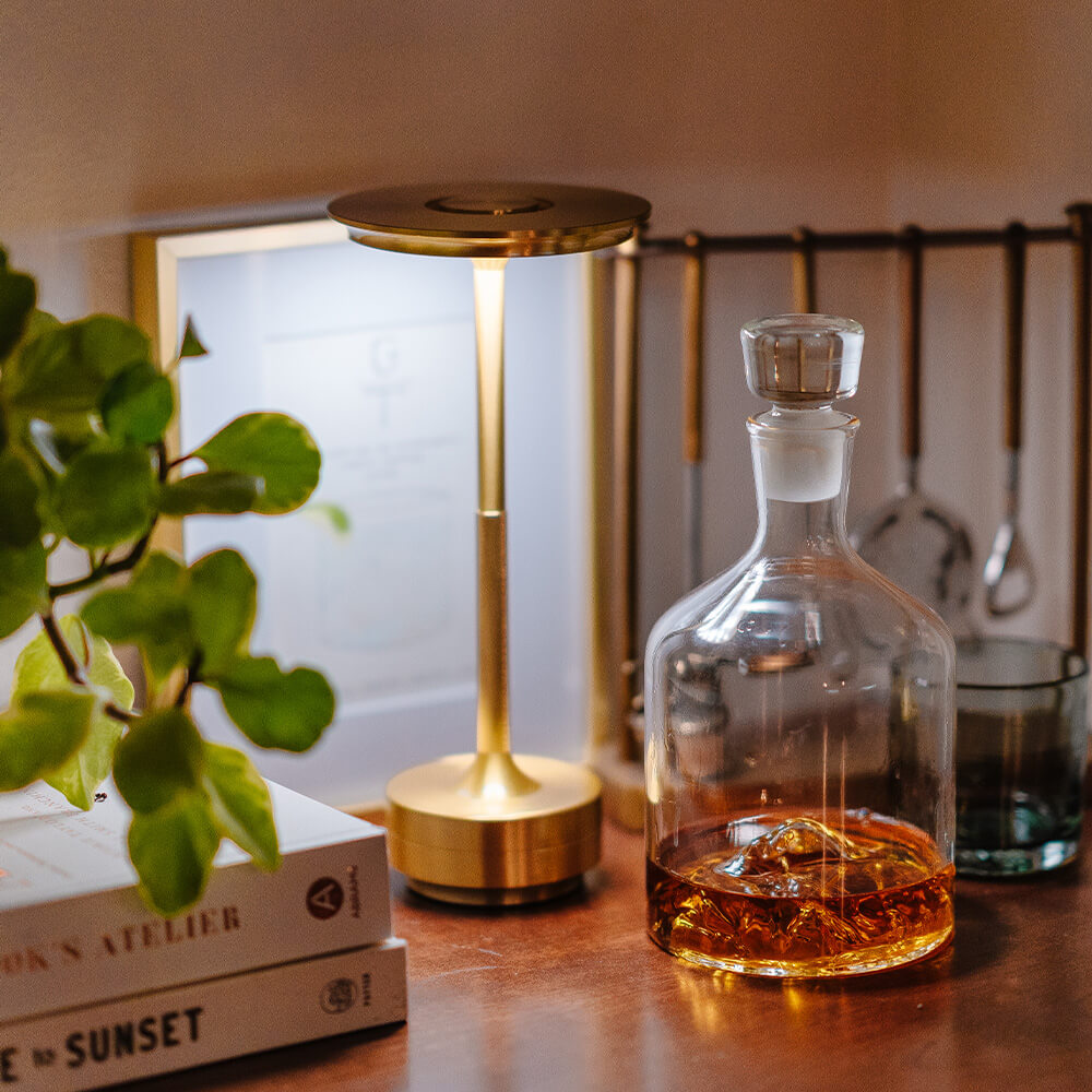 Brass cordless table lamp on a bar table beside a glass decanter and books, creating a cozy atmosphere.