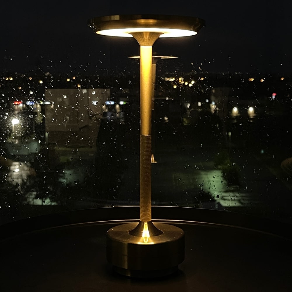 Gold cordless table lamp illuminated at night with a rainy cityscape in the background.