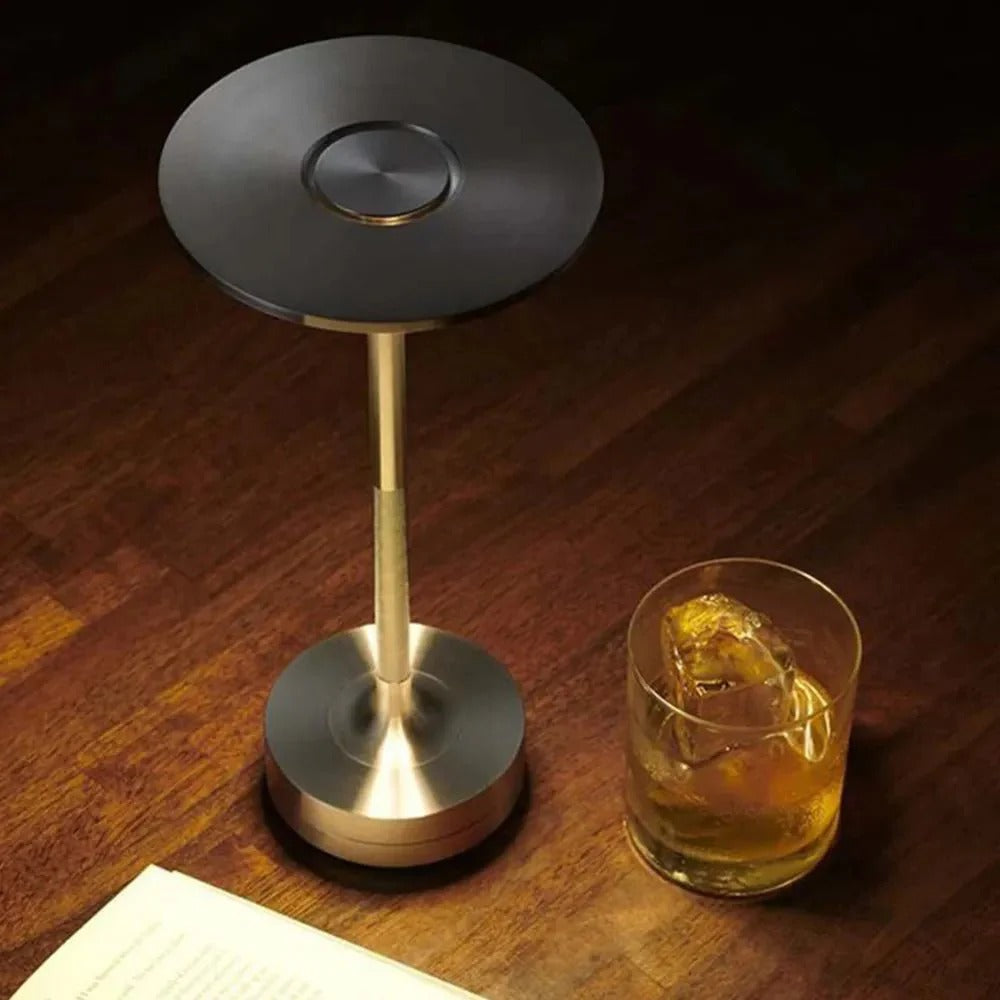 Brass cordless table lamp on a wooden surface, next to a glass of whiskey, providing ambient lighting.