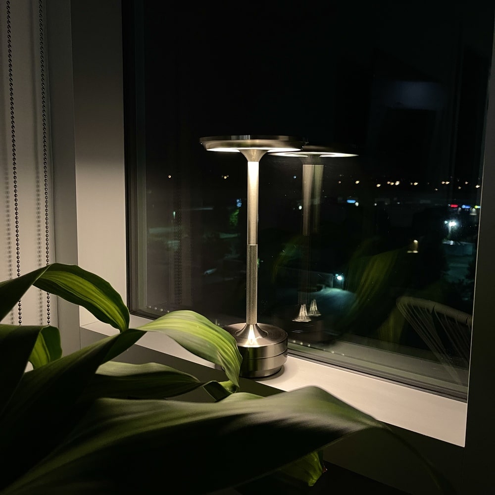 Silver rechargeable table lamp glowing in the dark, placed on a windowsill next to a plant.