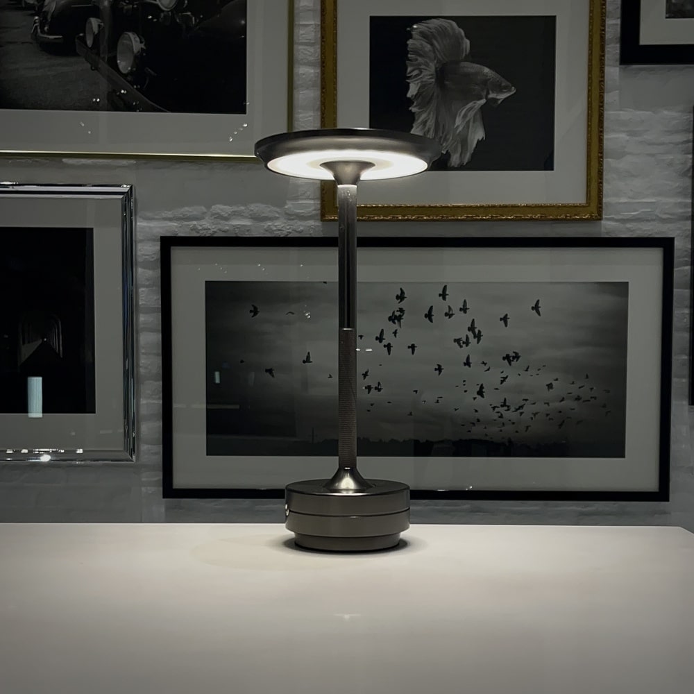 Modern cordless table lamp in a sleek silver design, illuminated against a backdrop of framed black and white artwork, ideal for home office or living room settings.