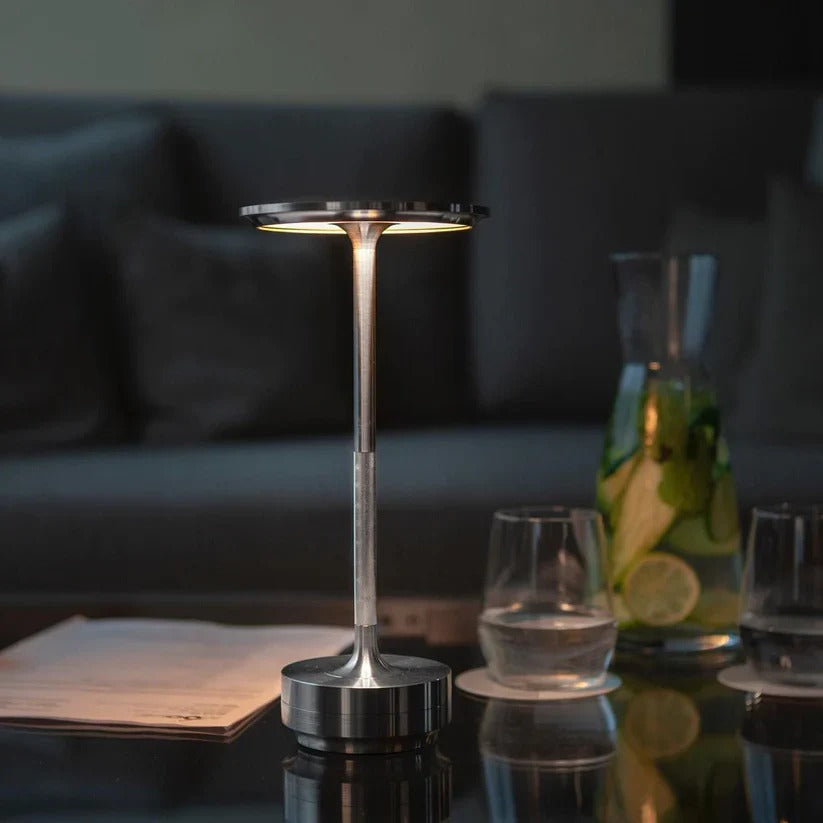 Rechargeable silver portable table lamp placed on a glass coffee table in a cozy living room, perfect for reading or creating a relaxing ambiance.