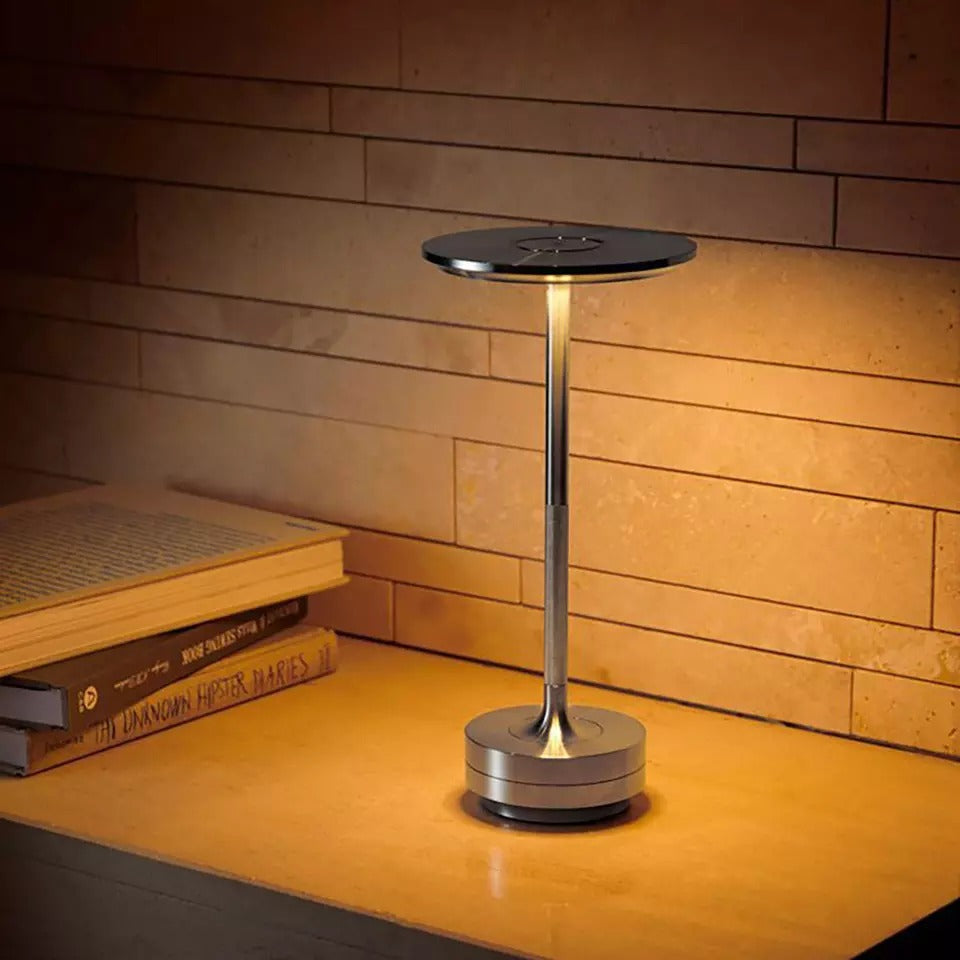 Stylish LED cordless table lamp with a metallic finish, providing warm lighting on a wooden desk next to books, ideal for workspaces or home offices.