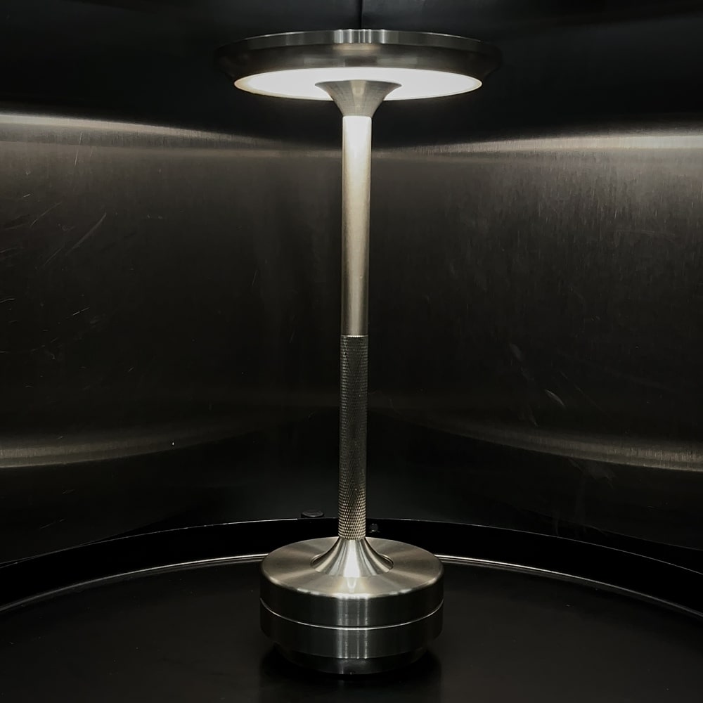 Rechargeable cordless table lamp in a metallic finish, displayed in a dark reflective environment.