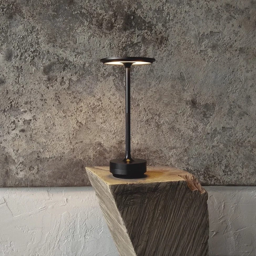 Cordless rechargeable table lamp on a textured wooden block against a rugged stone wall