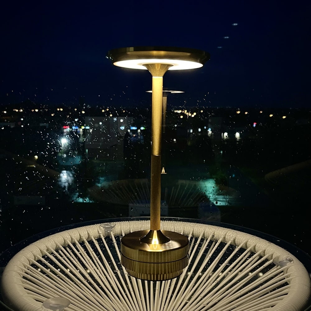 Gold cordless table lamp with LED lighting, placed on a white rope table during a rainy night.