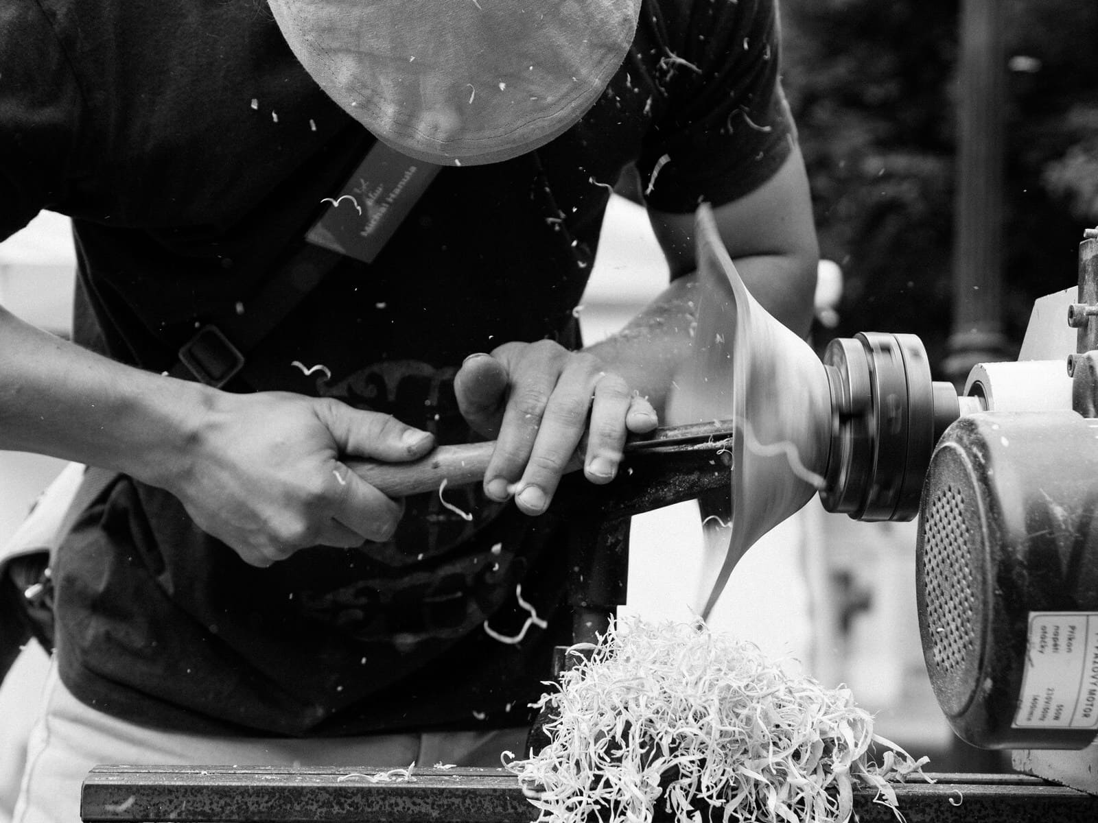 A black-and-white image of an artisan shaping custom lighting fixtures on a lathe, surrounded by wood shavings. The scene highlights traditional craftsmanship and attention to detail in custom lighting production.