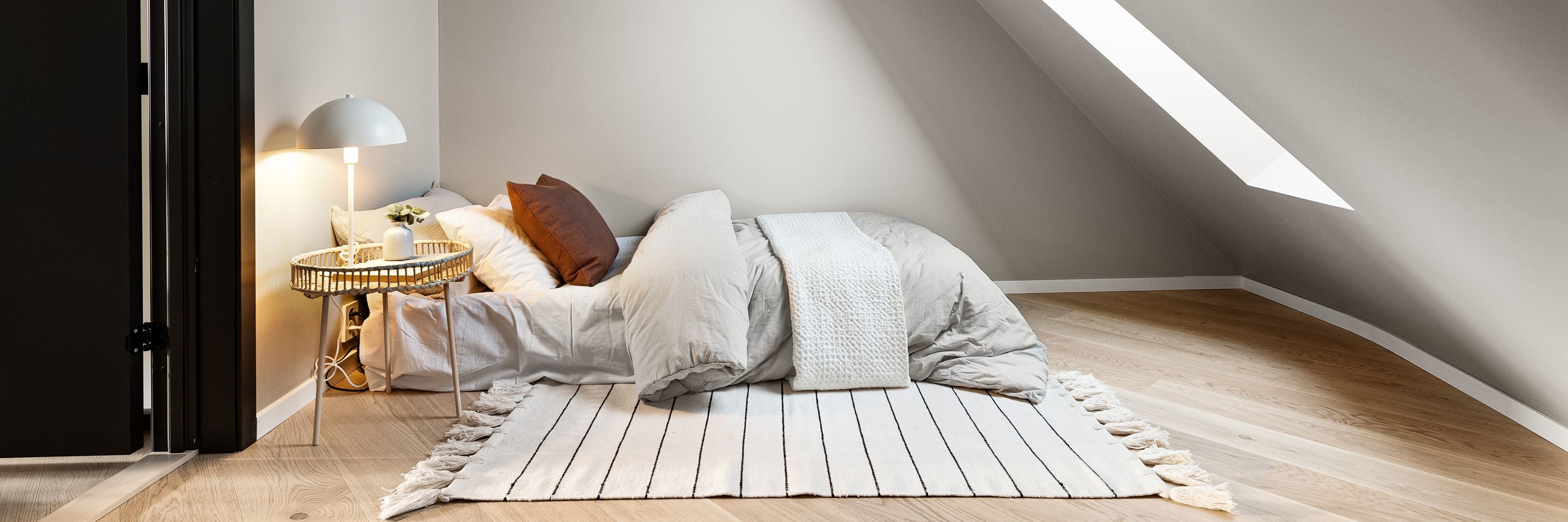 A cozy attic bedroom with a modern table lamp on a side table, illuminating a bed with warm pillows and a striped rug.