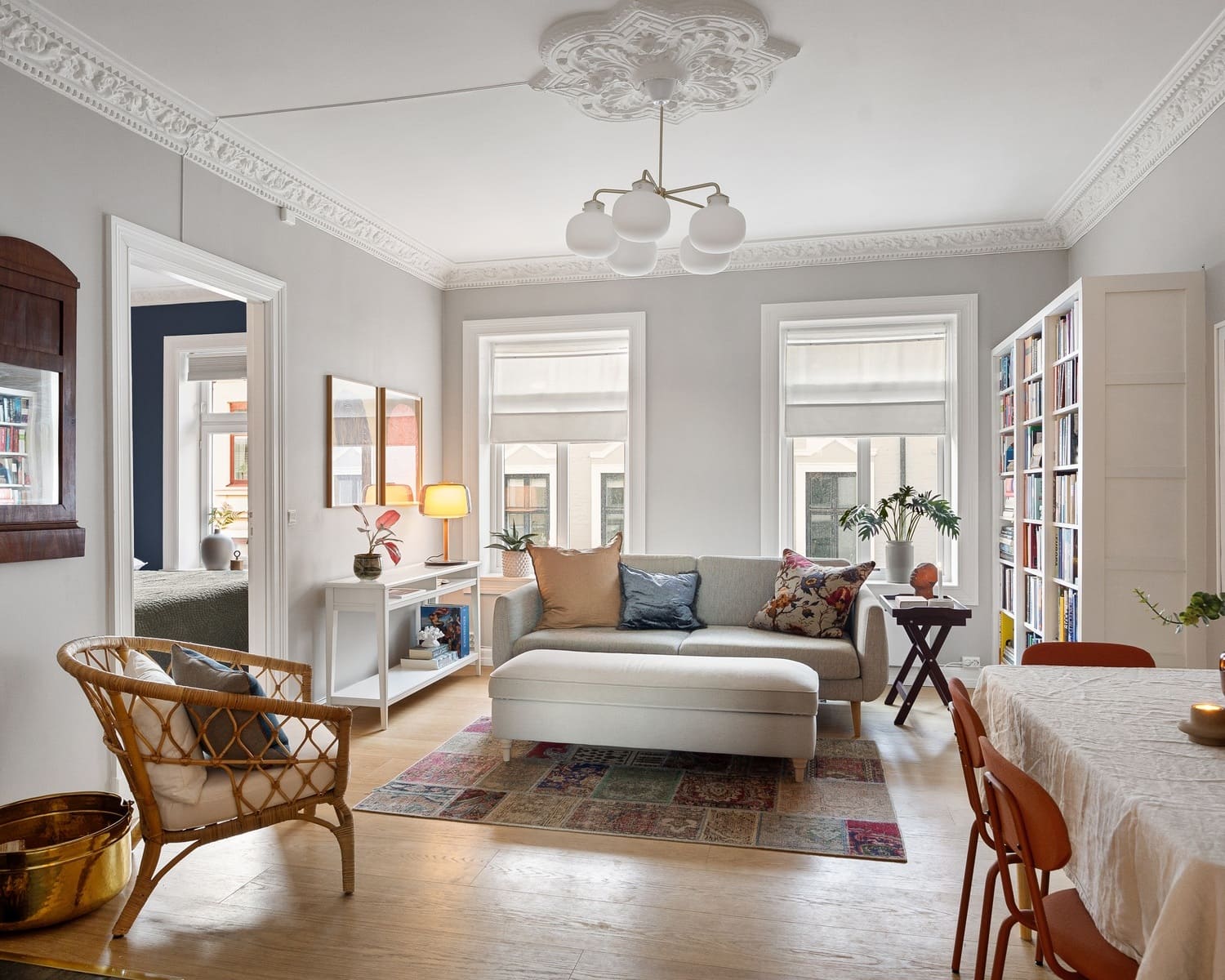 A vintage-inspired living room with a classic chandelier featuring multiple globes. The room includes a comfortable sofa, a woven chair, and a mixture of modern and vintage decorations like a colorful rug and a small dining area.