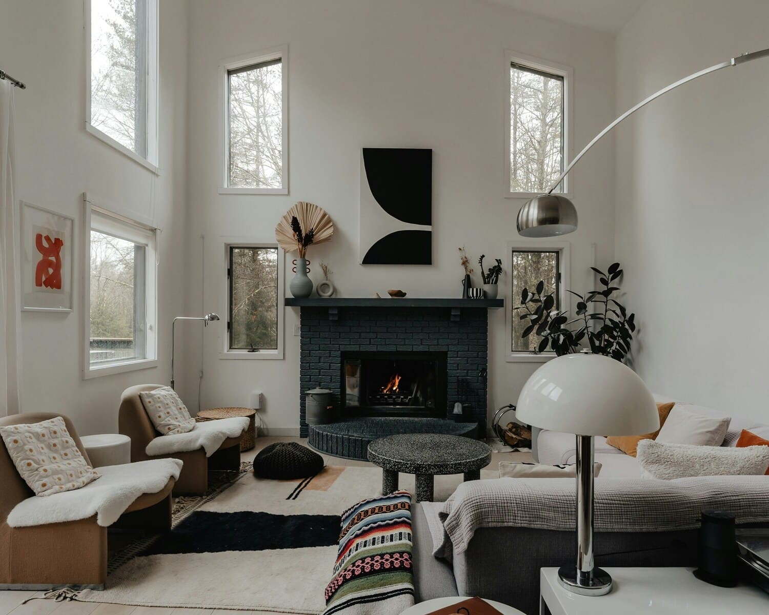 A contemporary living room with high ceilings, multiple windows, a black brick fireplace, and a combination of floor lamps and table lamps, complemented by a mix of neutral and warm tones in the furniture and decor.