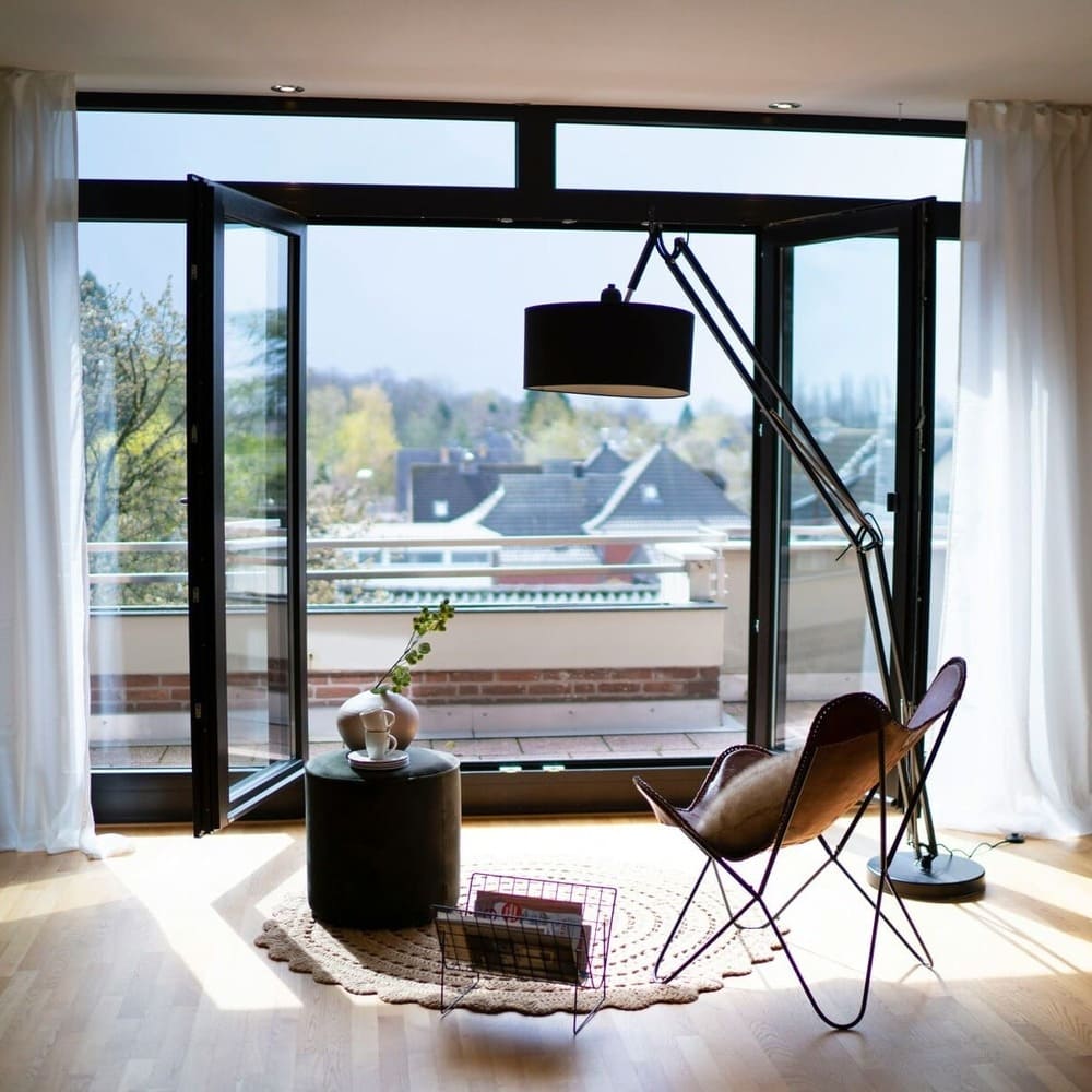 A cozy balcony setup with large open doors, a butterfly chair, a floor lamp, and a small round side table with a plant and vase. The space is flooded with natural light and offers a beautiful view of the surrounding neighborhood.