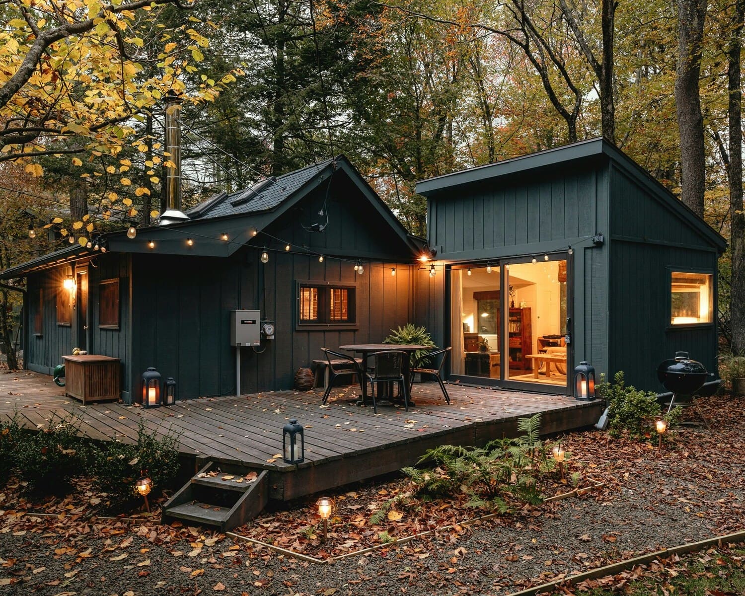 An inviting cabin exterior with dark wood paneling, a spacious deck adorned with lanterns and string lights, set against a backdrop of a forest in autumn, creating a warm and cozy ambiance.