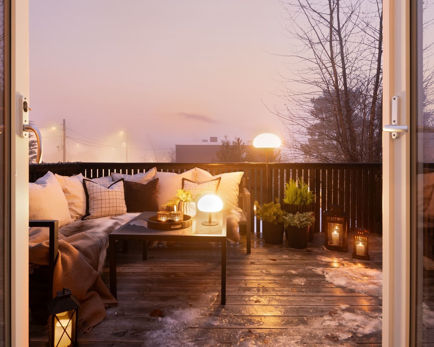 A cozy outdoor patio setup at dusk with warm lighting from table lamps and lanterns, a comfortable sofa with cushions, and potted plants, creating a relaxing and inviting atmosphere.