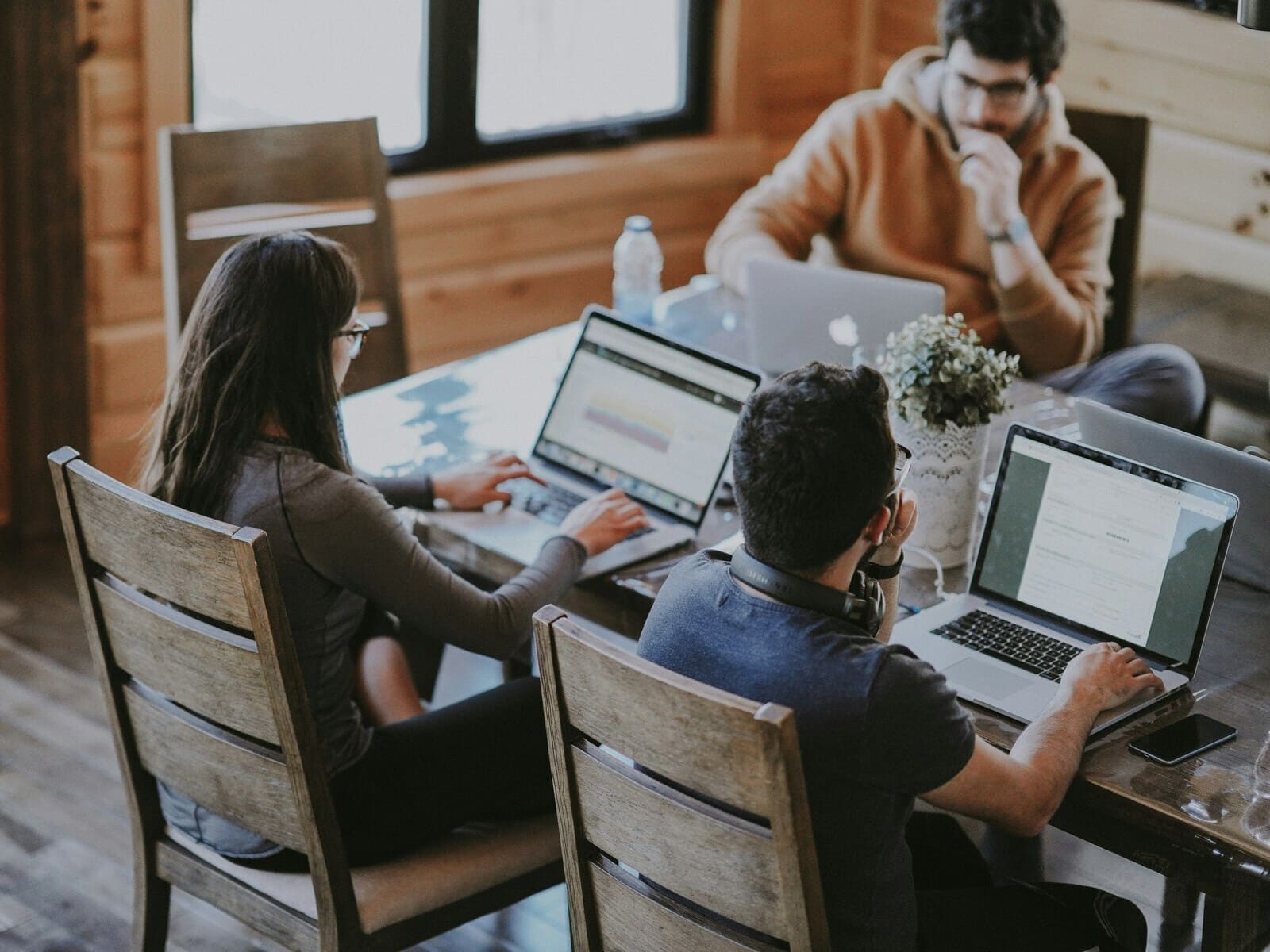 A customer support team working in a modern office with wooden interiors. Three individuals are seated around a table with laptops, focused on their tasks in a collaborative environment.