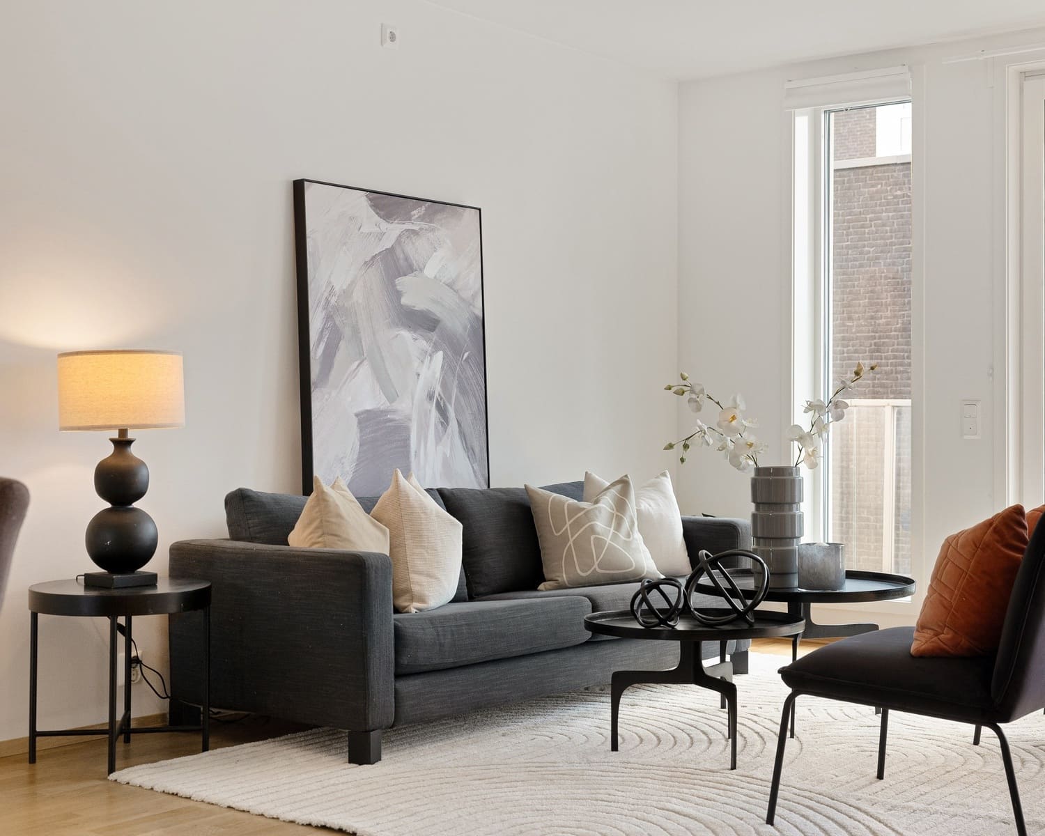 A contemporary living room with a dark gray sofa, a black side table with a spherical base table lamp, abstract wall art, and minimalist decor elements, including a modern vase with white flowers.