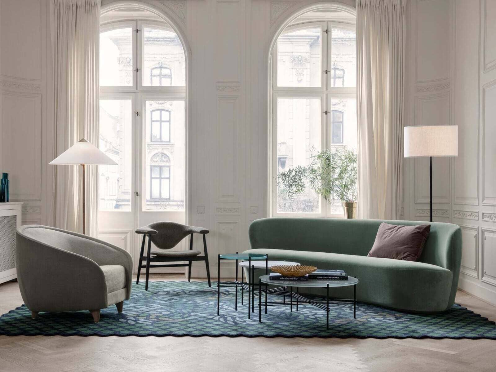 An elegant living room featuring large arched windows, a green sofa, a gray armchair, a stylish black chair, and layered round coffee tables on a blue patterned rug. The room is illuminated by floor lamps and natural light from the tall windows.