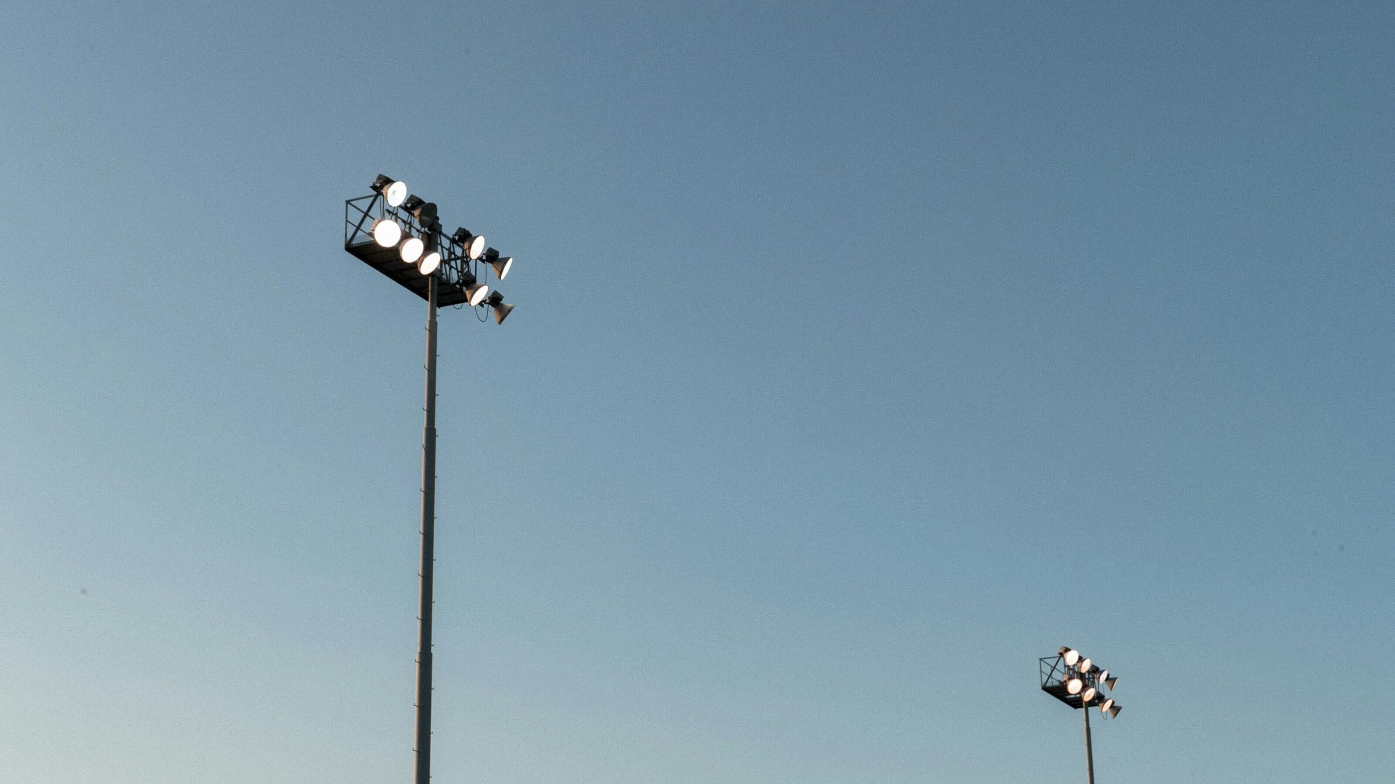 Outdoor floodlights mounted on tall poles against a clear blue sky, creating a minimalistic and open setting. Ideal for use as a banner image on a FAQ page.