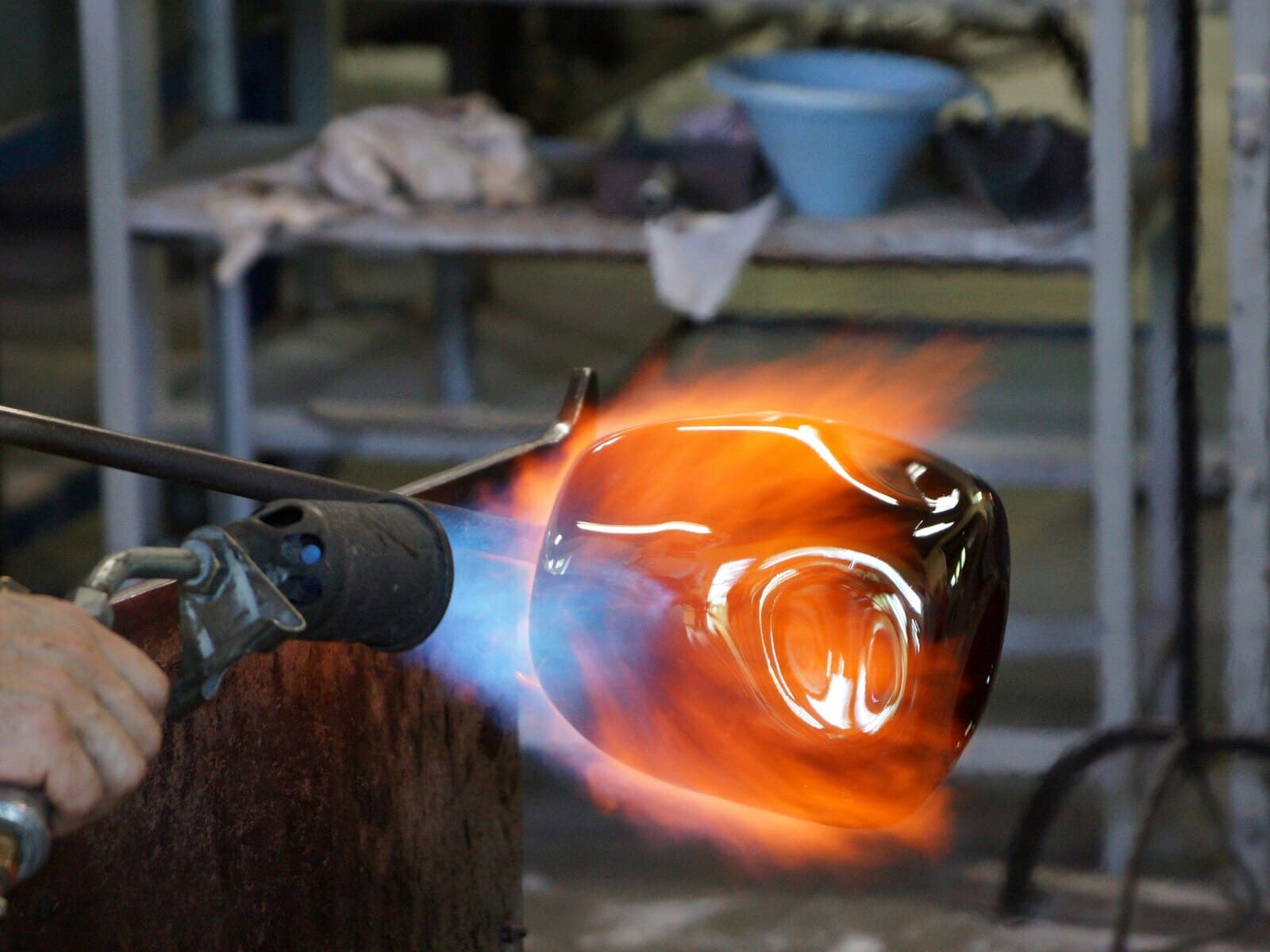 A close-up of a glass-blowing process for B2B custom lighting. A glowing piece of molten glass is being shaped with a torch, showcasing the craftsmanship and precision of the artisan.