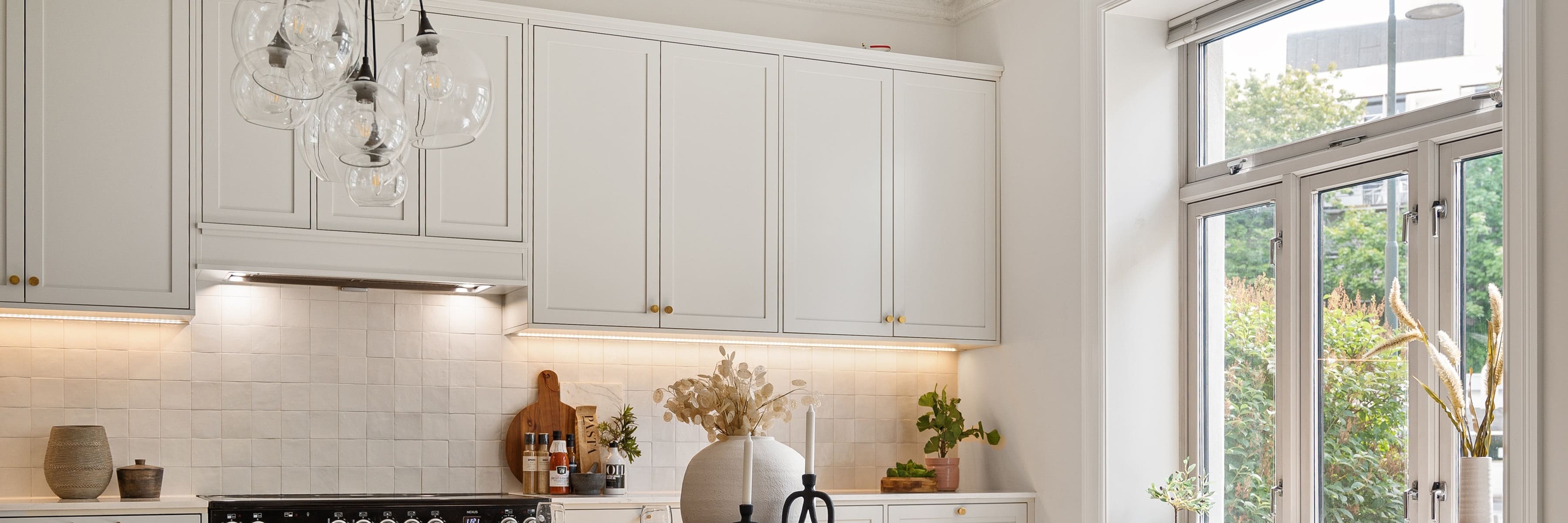 Modern kitchen with white cabinets and a stylish glass bulb chandelier illuminating the space. A minimalist design featuring natural light from large windows and a vase with dried flowers on the countertop.