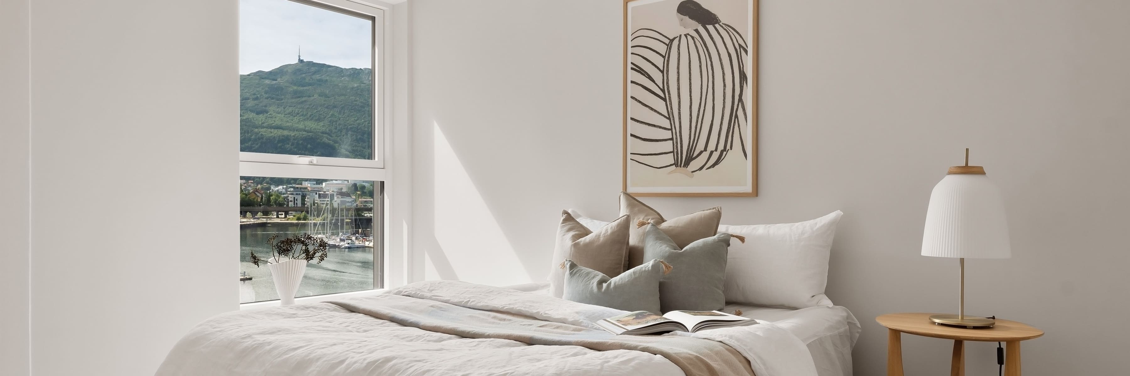 A Scandinavian-style bedroom with a wooden side table featuring a minimalist table lamp with a white pleated shade, neutral-colored bedding, decorative pillows, and a large window with a view of a hillside.