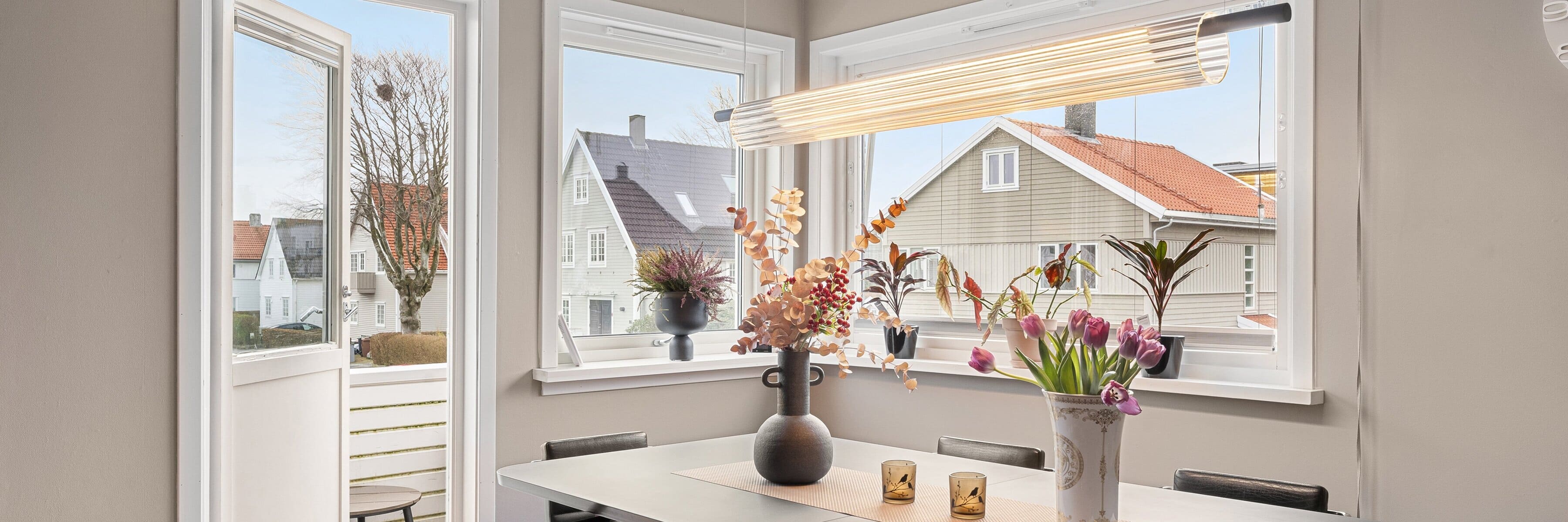 A modern dining room with large windows, decorated with a geometric pendant light, minimalist table setting, and fresh flowers.