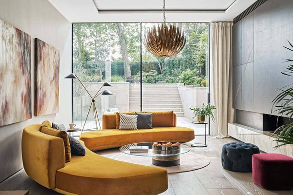 A modern living room featuring a mustard yellow curved sofa, large glass windows overlooking a garden, and stylish decor including abstract paintings, a unique chandelier, and a round coffee table. The space is filled with natural light and greenery.