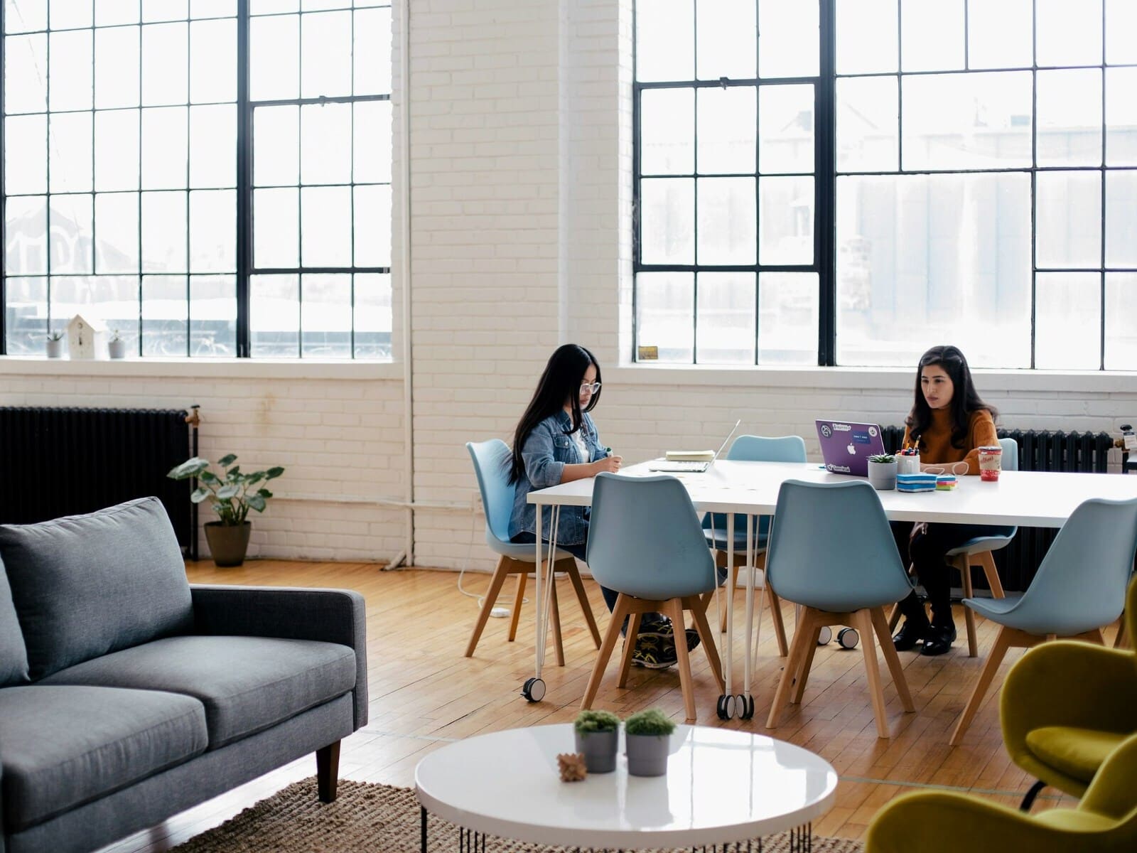 A modern office space with large industrial-style windows, featuring two women working at a white table with light blue chairs. The room has a gray sofa, light wood flooring, and minimalistic decor, creating a collaborative and bright atmosphere.